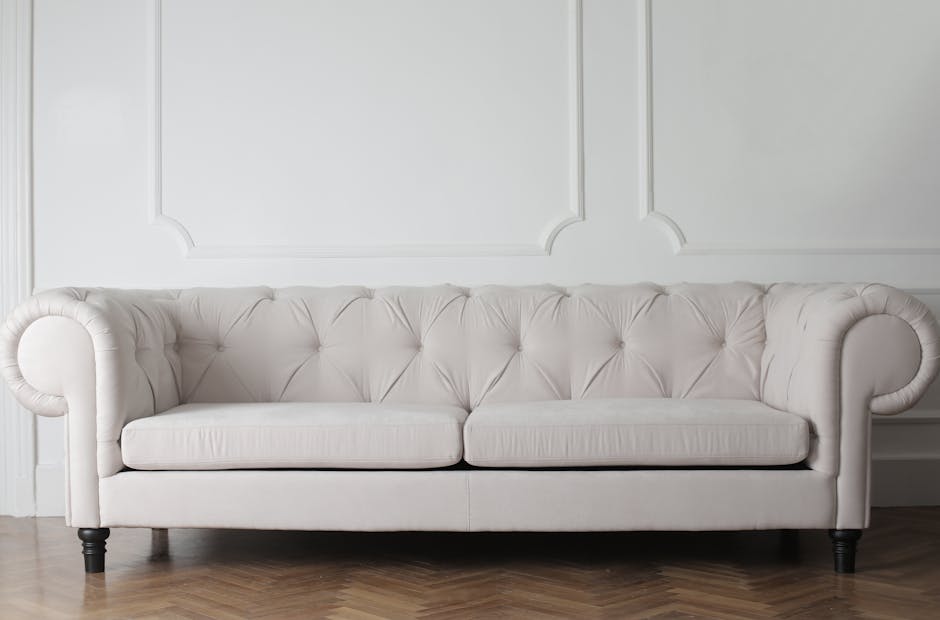 A white, tufted fabric sofa with rolled arms and dark wooden legs positioned against a paneled white wall in a well-lit room. The sofa surface appears clean and free of dust or stains, reflecting prof